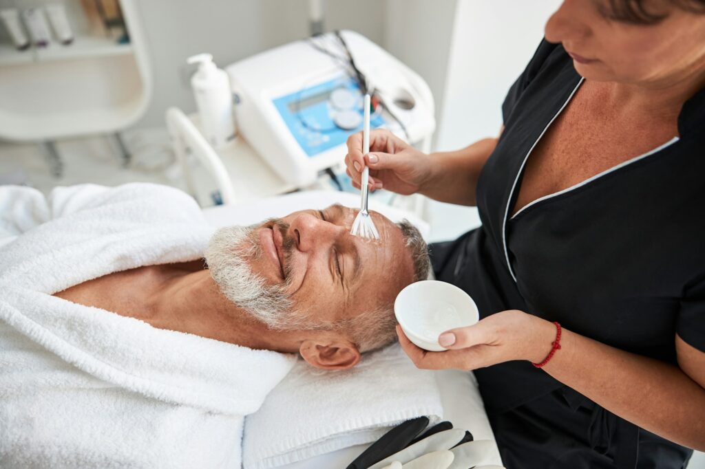 Skillful cosmetology specialist putting lotion on face of a man